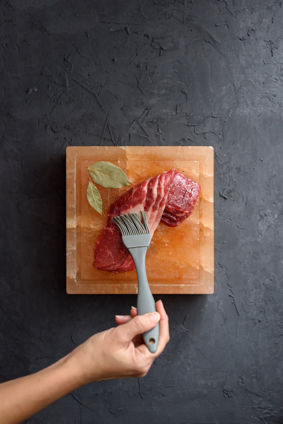 Salt block cooking concept. Woman hand brushing raw meat with oil on Himalayan pink salt block on black background copy space